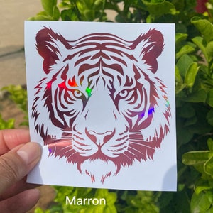 May include: A square white decal featuring a stylized tiger head graphic in a deep maroon color with a holographic finish that reflects rainbow light. The tiger design displays intricate facial stripes and sharp, focused eyes on a white background.