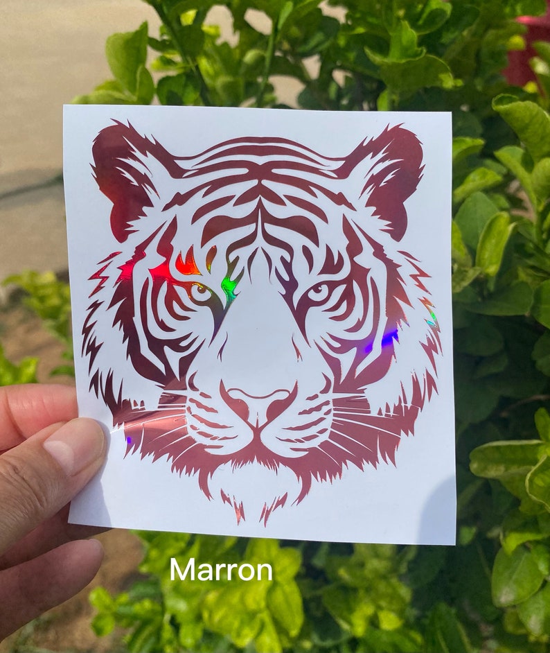 May include: A square white decal featuring a stylized tiger head graphic in a deep maroon color with a holographic finish that reflects rainbow light. The tiger design displays intricate facial stripes and sharp, focused eyes on a white background.
