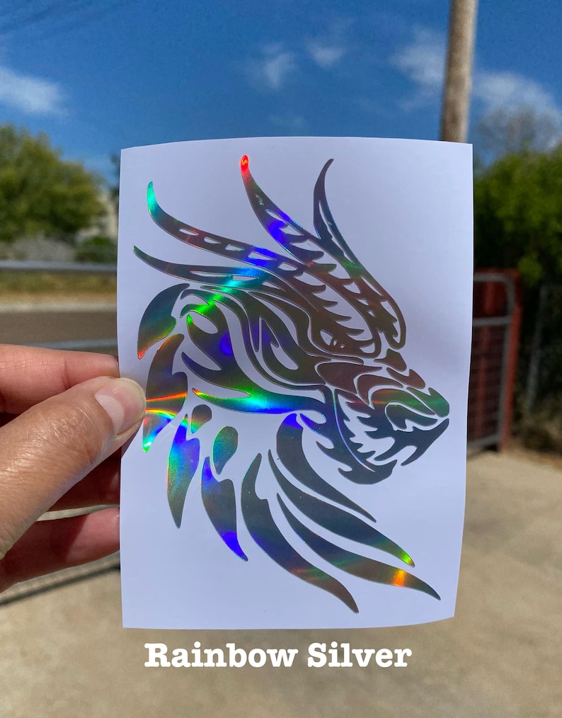 K&ouml;nnte beinhalten: Ein silberner, schimmernder Vinyl-Aufkleber mit einem Drachenkopf mit Tribal-Design. Der Aufkleber befindet sich auf einem wei&szlig;en Hintergrund. Rainbow Silver.