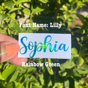 May include: A rectangular white sticker featuring the name Sophia in a cursive script font. The lettering displays a holographic rainbow green finish. Text printed on the image includes Font Name: Lilly and Rainbow Green.