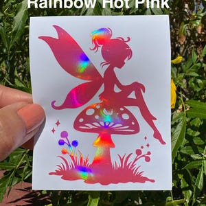 May include: A pink sticker featuring a fairy silhouette with rainbow holographic wings and accents, seated on a mushroom. The sticker has the text "Rainbow Hot Pink" at the top. The fairy is surrounded by stars and grass.