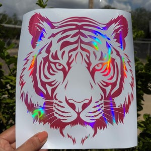 May include: A decal featuring a stylized tiger head graphic in hot pink with a holographic rainbow finish. The design is printed on a white rectangular backing sheet. The text "Rainbow hot pink" is printed in white at the top of the image.