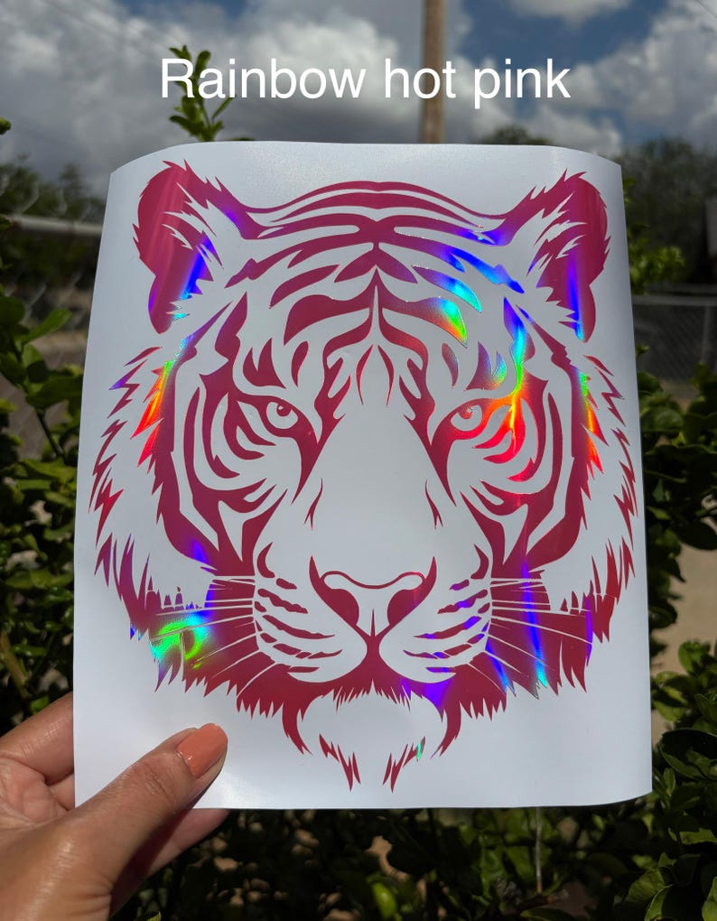 May include: A decal featuring a stylized tiger head graphic in hot pink with a holographic rainbow finish. The design is printed on a white rectangular backing sheet. The text "Rainbow hot pink" is printed in white at the top of the image.