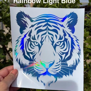 May include: A square decal featuring a stylized tiger head graphic with a rainbow light blue holographic finish. The design consists of intricate blue and iridescent lines forming the tiger's face, set against a plain white backing sheet.