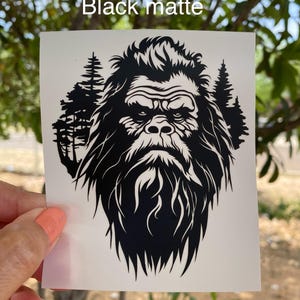 May include: A black matte vinyl decal featuring a detailed graphic illustration of a Bigfoot face centered against a backdrop of silhouetted pine trees. The text Black matte is printed in white at the top of the square white backing sheet.