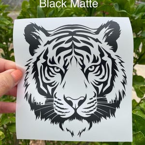 May include: A black matte vinyl decal featuring a detailed graphic illustration of a tiger face. The design is printed on a white square backing sheet with the text Black Matte visible at the top of the image, showcasing sharp lines.