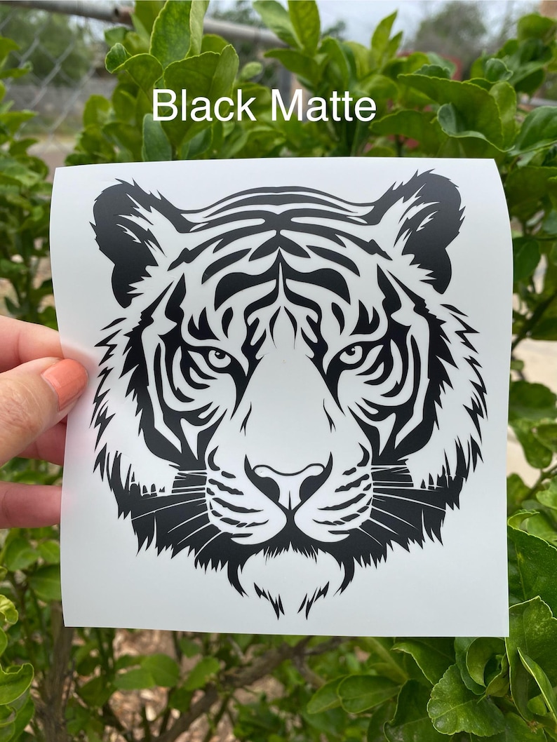 May include: A black matte vinyl decal featuring a detailed graphic illustration of a tiger face. The design is printed on a white square backing sheet with the text Black Matte visible at the top of the image, showcasing sharp lines.