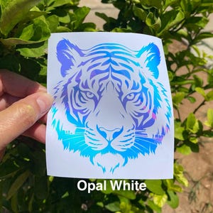 May include: A square decal featuring a stylized tiger head graphic with an iridescent opal white finish that reflects blue and purple hues. The design is printed on a white backing, and the text Opal White is printed below the tiger graphic.