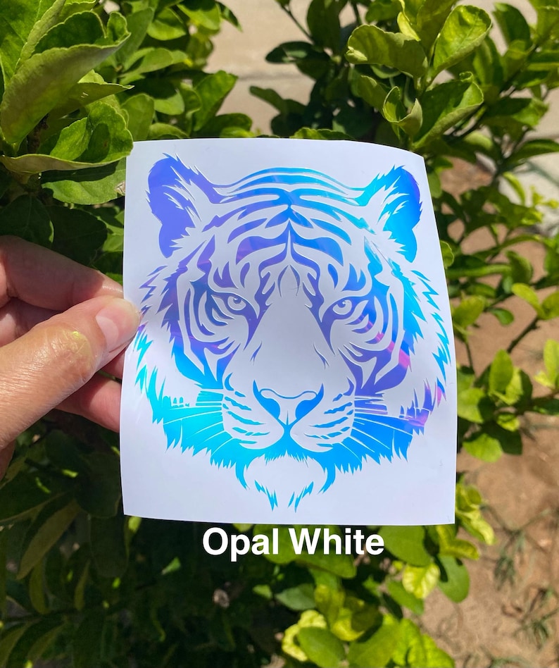 May include: A square decal featuring a stylized tiger head graphic with an iridescent opal white finish that reflects blue and purple hues. The design is printed on a white backing, and the text Opal White is printed below the tiger graphic.