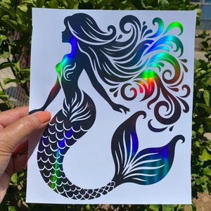 K&ouml;nnte beinhalten: Schwarzer Meerjungfrau-Aufkleber mit holografischen Regenbogenakzenten. Der Aufkleber tr&auml;gt oben den Text "Rainbow Black". Die Meerjungfrau hat lange, flie&szlig;ende Haare und einen detaillierten Schwanz. Der Aufkleber wird von einer Hand gehalten.