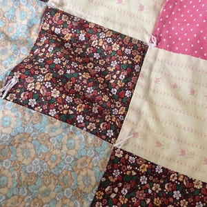 May include: Close-up of a patchwork quilt with various floral and patterned fabrics. The quilt features squares of burgundy floral, light pink polka dots, and cream-colored fabric with a tulip design. The quilt is hand-stitched.
