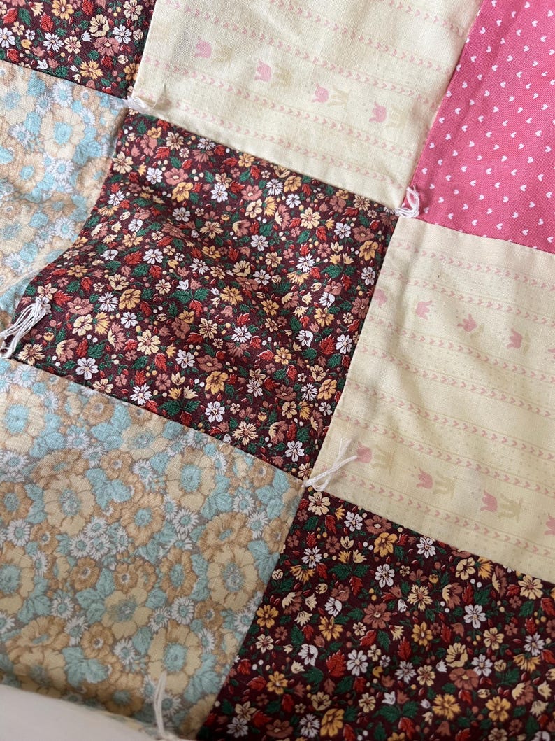 May include: Close-up of a patchwork quilt with various floral and patterned fabrics. The quilt features squares of burgundy floral, light pink polka dots, and cream-colored fabric with a tulip design. The quilt is hand-stitched.
