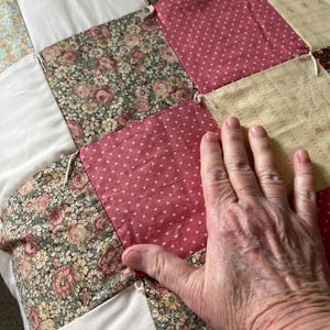 May include: Close-up of a patchwork quilt with various floral and dotted patterns in shades of pink, cream, and green. The quilt is made of square fabric pieces sewn together. A hand rests on the quilt.
