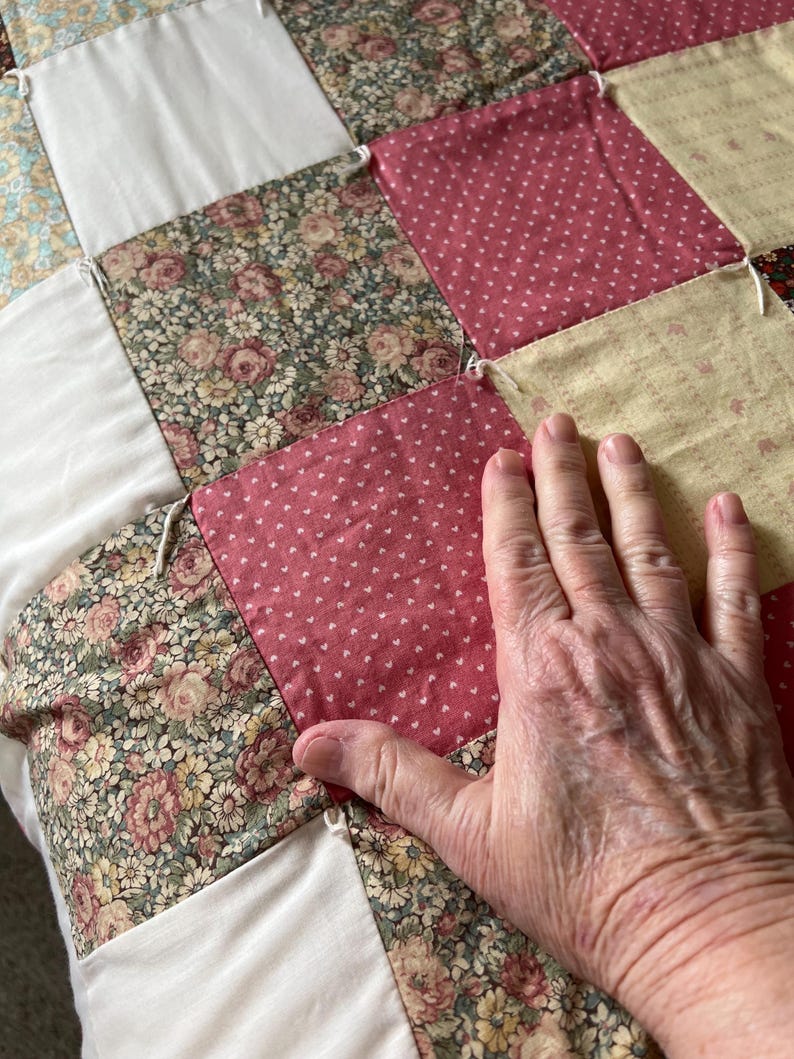 May include: Close-up of a patchwork quilt with various floral and dotted patterns in shades of pink, cream, and green. The quilt is made of square fabric pieces sewn together. A hand rests on the quilt.