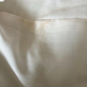 May include: Close-up of a section of off-white fabric, possibly linen or a similar material. The fabric has a subtle sheen and visible texture. There are faint orange-brown stains near the seam, adding a touch of color contrast to the neutral background.