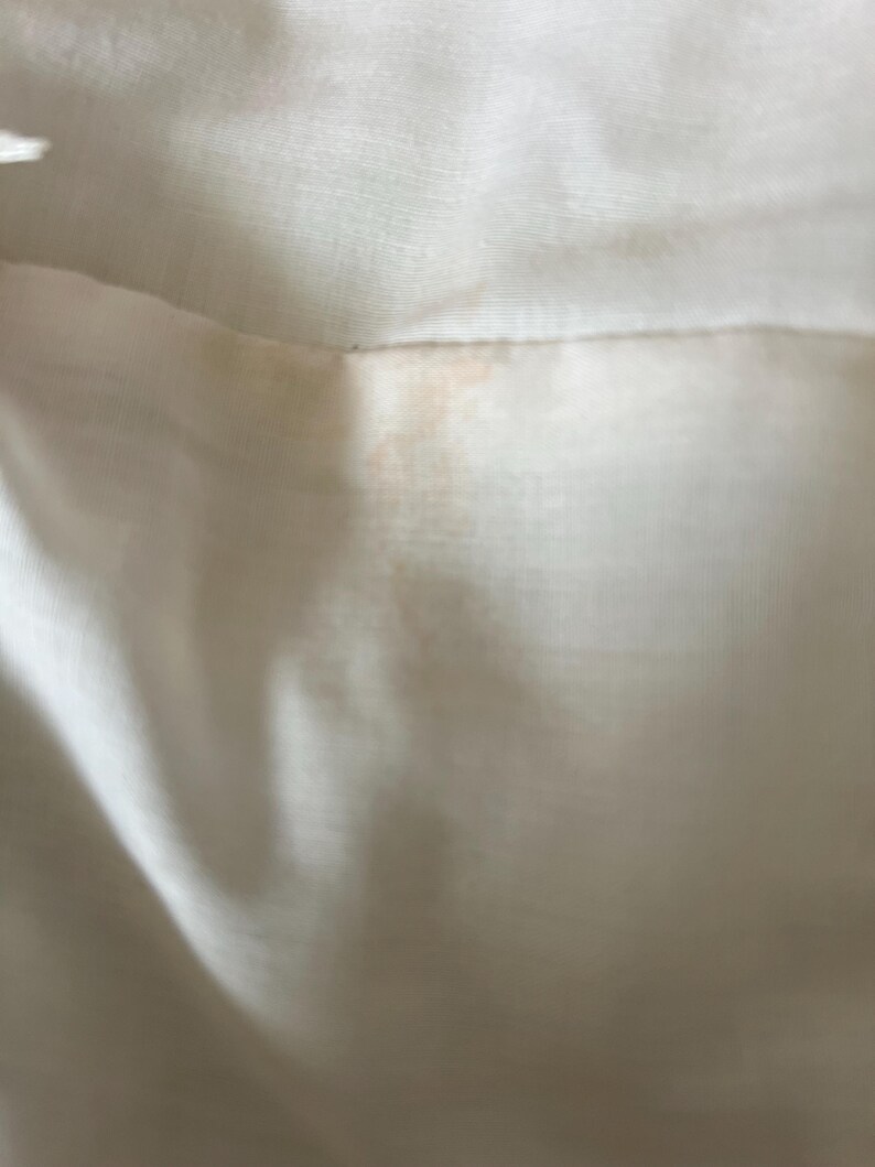May include: Close-up of a section of off-white fabric, possibly linen or a similar material. The fabric has a subtle sheen and visible texture. There are faint orange-brown stains near the seam, adding a touch of color contrast to the neutral background.