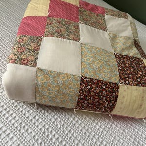 May include: A patchwork pillow with a variety of floral and polka dot patterns in pink, yellow, and brown. The pillow is made of squares sewn together, creating a textured surface. The pillow is resting on a white bedspread.
