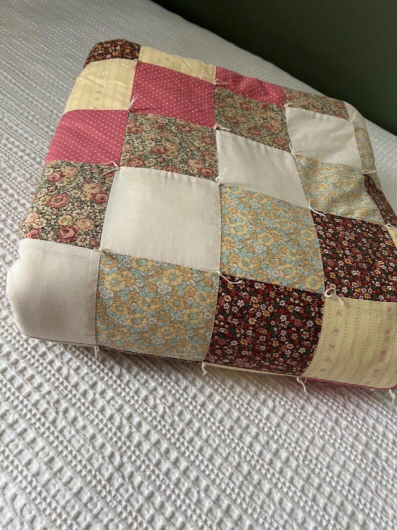 May include: A patchwork pillow with a variety of floral and polka dot patterns in pink, yellow, and brown. The pillow is made of squares sewn together, creating a textured surface. The pillow is resting on a white bedspread.