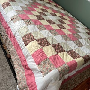 May include: A handmade quilt with a patchwork design in shades of pink, white, cream, and floral patterns. The quilt is on a bed with a dark metal frame. The quilt's border is a pink polka dot fabric.
