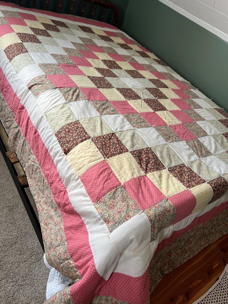May include: A handmade quilt with a patchwork design in shades of pink, white, cream, and floral patterns. The quilt is on a bed with a dark metal frame. The quilt's border is a pink polka dot fabric.