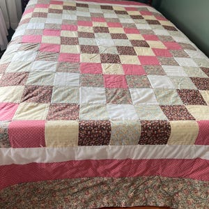May include: A patchwork quilt with a grid pattern of squares in various colors and patterns. The quilt features pink, white, yellow, and brown squares, with floral and polka dot designs. The bed skirt has a floral pattern.