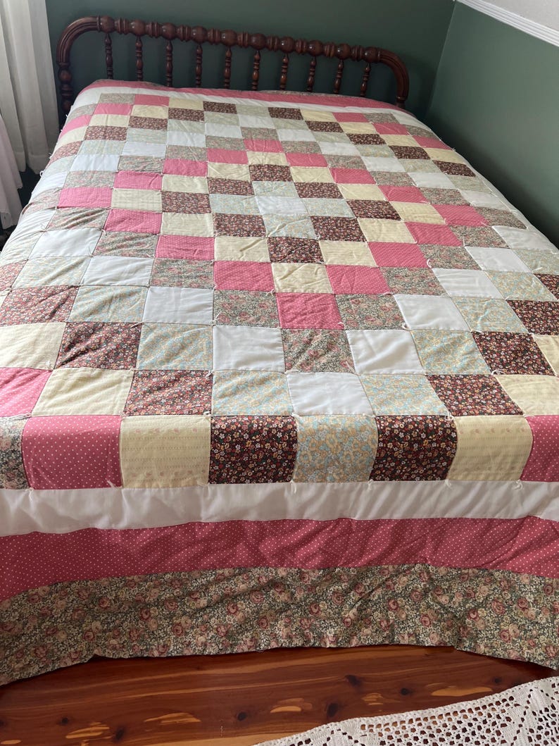 May include: A patchwork quilt with a grid pattern of squares in various colors and patterns. The quilt features pink, white, yellow, and brown squares, with floral and polka dot designs. The bed skirt has a floral pattern.