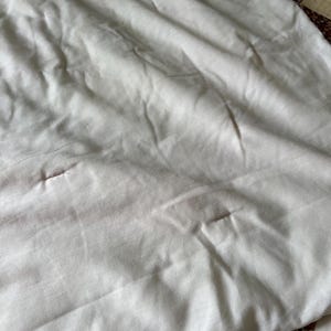 May include: Close-up of a white, slightly wrinkled fabric, possibly a sheet or blanket. The fabric has subtle variations in tone and texture, with a few small imperfections. A portion of a patchwork quilt with floral patterns is visible in the corner.