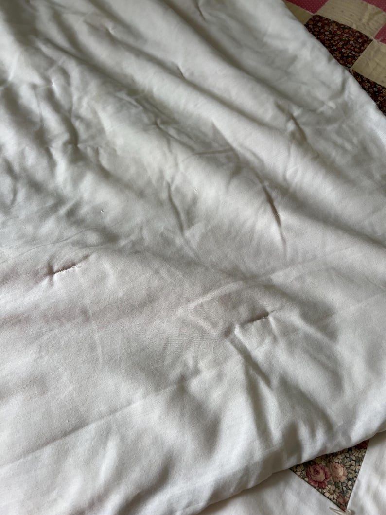 May include: Close-up of a white, slightly wrinkled fabric, possibly a sheet or blanket. The fabric has subtle variations in tone and texture, with a few small imperfections. A portion of a patchwork quilt with floral patterns is visible in the corner.