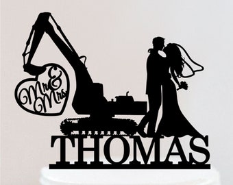 May include: Black silhouette of a couple kissing in front of an excavator with a heart that says "Mr. & Mrs." and the name "THOMAS" underneath. The couple is holding a bouquet of flowers. The silhouette is on a white cake.