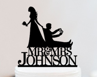 May include: Black silhouette cake topper of a bride pulling a groom who is holding a video game controller. The topper says "Mr. & Mrs. Johnson" in black lettering.