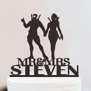 May include: A black cake topper with silhouettes of a couple holding hands, styled after comic book characters. The text "MR & MRS STEVEN" is below. The cake is white with floral decorations.