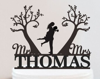 May include: Black silhouette cake topper featuring a couple embracing under two stylised trees with a heart shape formed by the branches. The text "Mr. & Mrs. THOMAS" is below the couple.