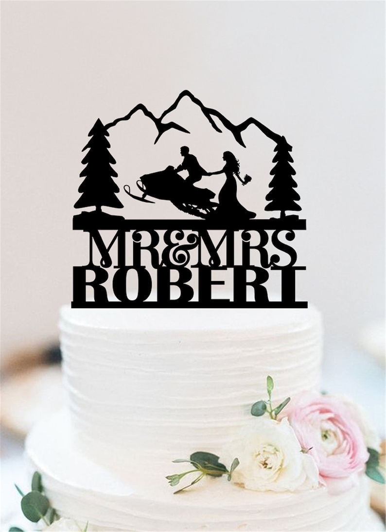 May include: Black silhouette cake topper of a couple riding a snowmobile in front of a mountain range with pine trees on either side. The text "MR & MRS ROBERT" is below the couple.
