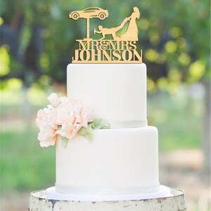 May include: A wooden wedding cake topper featuring a bride pulling a groom from under a car on a lift. The topper says "Mr. & Mrs. Johnson".