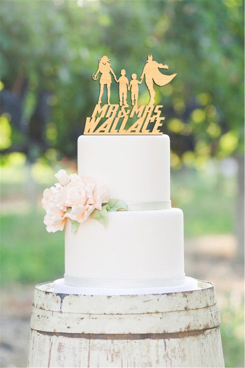 Może przedstawiać: Dwupiętrowy biały tort weselny z drewnianą nakładką przedstawiającą sylwetkę rodziny i napisem "MR & MRS WALLACE". Tort jest ozdobiony kompozycją kwiatową i stoi na rustykalnej beczce.
