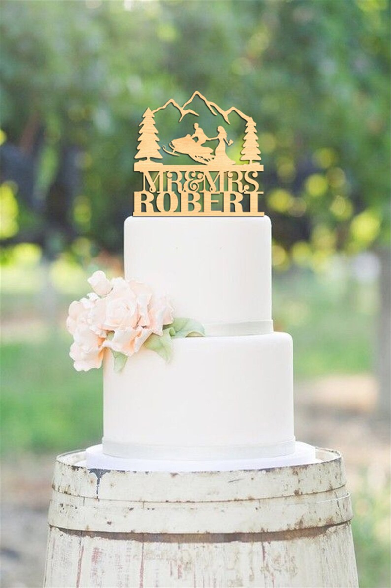 May include: A wooden wedding cake topper featuring a couple riding a snowmobile in front of a mountain range. The topper says "Mr. & Mrs. Robert".