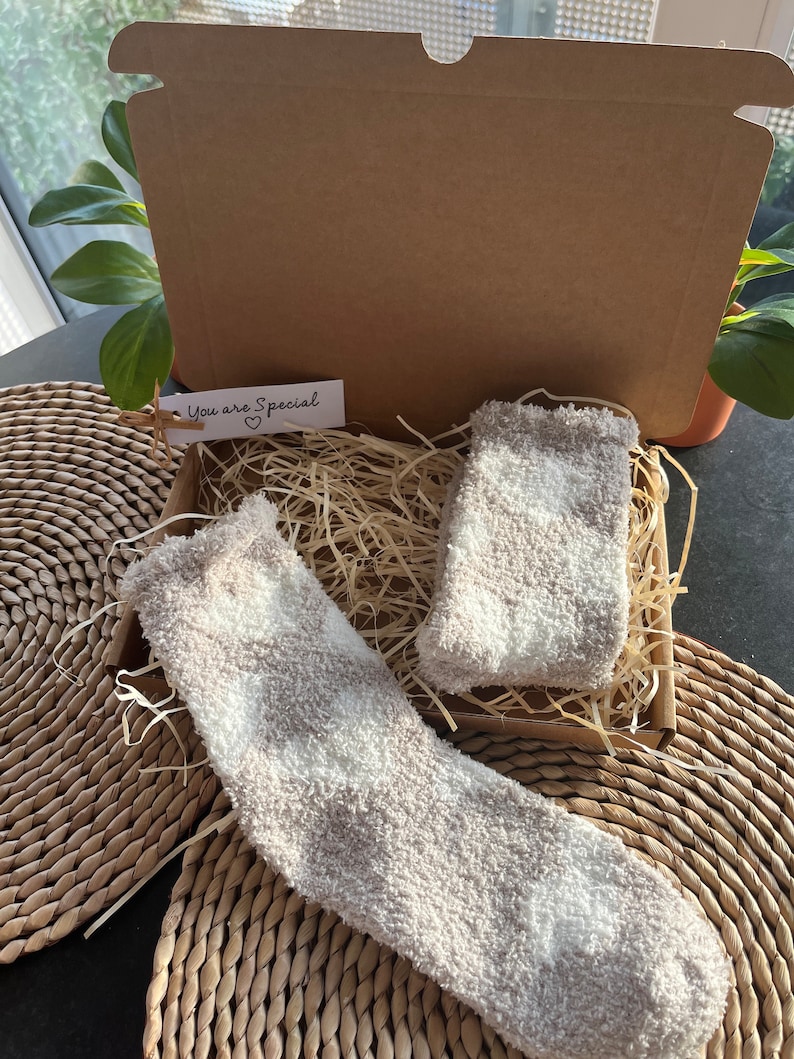 Pu&ograve; includere: Due paia di calzini morbidi, bianchi e grigi, in una scatola di cartone con carta triturata. La scatola ha un'etichetta che dice "You are Special" con un cuore.