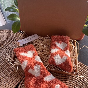 Pu&ograve; includere: Un paio di calzini morbidi con cuori bianchi su uno sfondo terracotta. I calzini sono confezionati in una scatola di cartone con carta triturata e un piccolo cartellino che dice "You are Special" con un cuore.