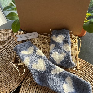 Pu&ograve; includere: Un paio di calzini blu e pelosi con motivi a forma di cuore bianchi in una scatola regalo con un'etichetta che dice "You are Special" con un simbolo di cuore.