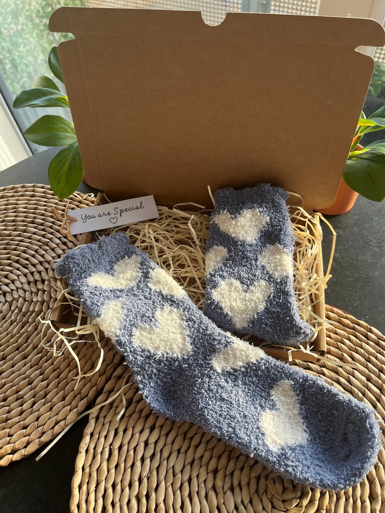 Pu&ograve; includere: Un paio di calzini blu e pelosi con motivi a forma di cuore bianchi in una scatola regalo con un'etichetta che dice "You are Special" con un simbolo di cuore.