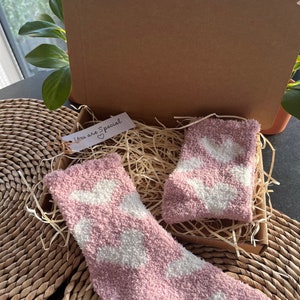 Pu&ograve; includere: Un paio di calzini rosa e pelosi con disegni di nuvole bianche sono in una scatola di cartone con carta triturata. La scatola ha un piccolo cartellino che dice "You are Special" con un cuore.