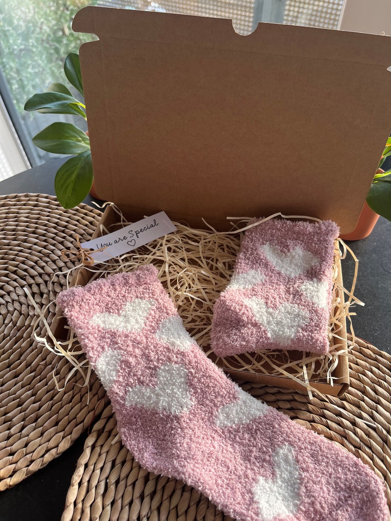 Pu&ograve; includere: Un paio di calzini rosa e pelosi con disegni di nuvole bianche sono in una scatola di cartone con carta triturata. La scatola ha un piccolo cartellino che dice "You are Special" con un cuore.