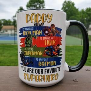 May include: White ceramic mug with a black handle. The mug has a colorful design featuring the text "Daddy You Are" and images of superheroes, including Iron Man, Hulk, Superman, and Batman. The text "You Are Our Favorite Superhero" is printed at the bottom of the mug.