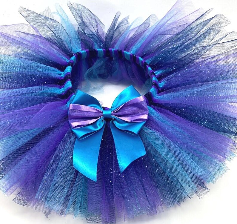 May include: A vibrant tutu skirt in shades of blue and purple, featuring layers of tulle. A large satin bow in turquoise and purple accents the front. The skirt is designed for children's wear.