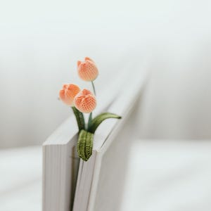 May include: A close-up of a book with a crocheted flower bookmark. The bookmark is made of pink and green yarn and features three small tulips.
