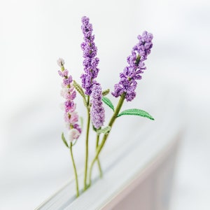 May include: A close-up of crocheted lavender sprigs in shades of purple and pink, with green stems and leaves, placed in a book. The handmade flowers have a textured appearance against a soft, white background.