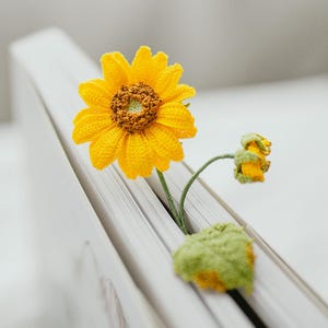 May include: A handmade, yellow crochet sunflower bookmark with a green stem and leaf, nestled between the pages of a white book. The sunflower has a detailed, textured center and petals, and a smaller bud is also visible.
