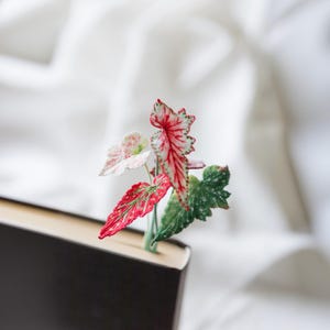 May include: A handmade bookmark featuring colorful fabric leaves in shades of red, green, and pink, inserted into a closed book. The leaves have intricate details and are set against a white background.