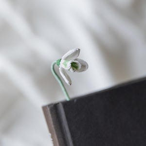 May include: A handmade crochet bookmark featuring a white snowdrop flower with green accents. The flower is attached to a green stem, emerging from the top of a dark book. The bookmark is a unique, handcrafted item for book lovers.