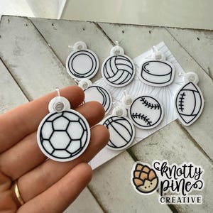 May include: A collection of white and black sports-themed charms, including a soccer ball, volleyball, hockey puck, baseball, basketball, and football. Each charm has a clear plastic loop for hanging. The image also includes a logo that says "knotty pines creative".